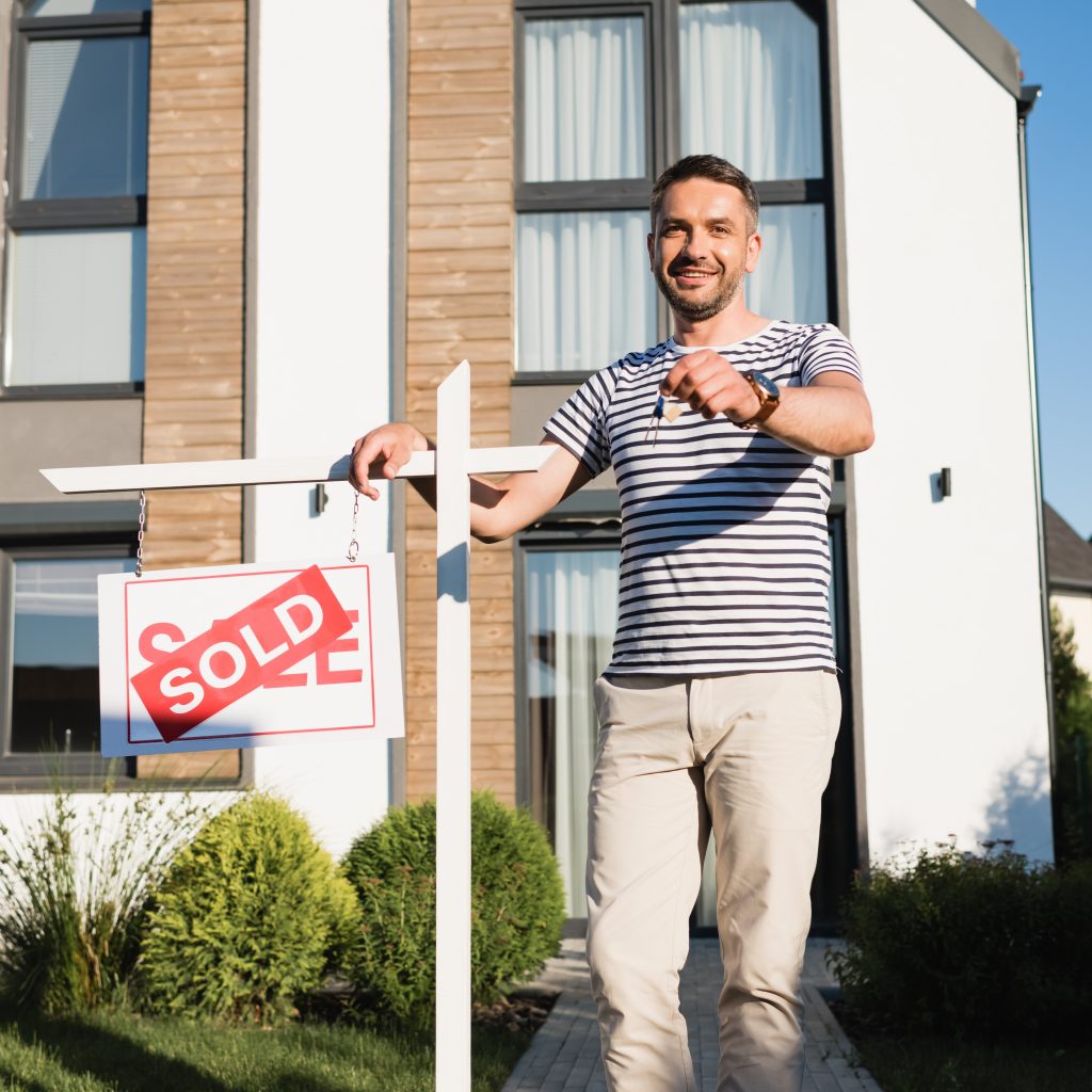 Full length of smiling man showing key while standing near sign with sold lettering with modern house on background Happy man holding keys in front of modern house with SOLD sign, celebrating a successful FSBO home sale